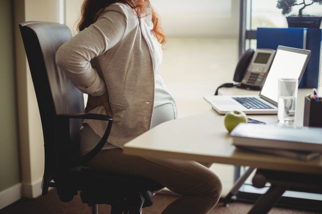 Pregnant woman at office desk holding her lower back in discomfort while working